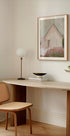 un espace de travail moderne et minimaliste avec un bureau en bois, une chaise tressée, une lampe et une peinture encadrée d'une maison.