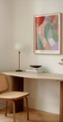 un espace de travail minimaliste avec un bureau en bois, une chaise tressée, une lampe et une peinture abstraite encadrée au mur.