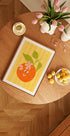 une table en bois avec une image encadrée d'une orange, un bol de citrons, un vase de tulipes roses et une bougie dessus.