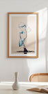 une œuvre d'art encadrée représentant une gouttelette d'eau suspendue dans les airs, avec une palette de couleurs bleu et blanc. L'œuvre est exposée sur un mur blanc au-dessus d'une table avec un vase blanc et un livre.