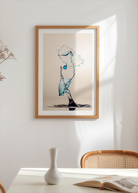 une œuvre d'art encadrée représentant une gouttelette d'eau suspendue dans les airs, avec une palette de couleurs bleu et blanc. L'œuvre est exposée sur un mur blanc au-dessus d'une table avec un vase blanc et un livre.