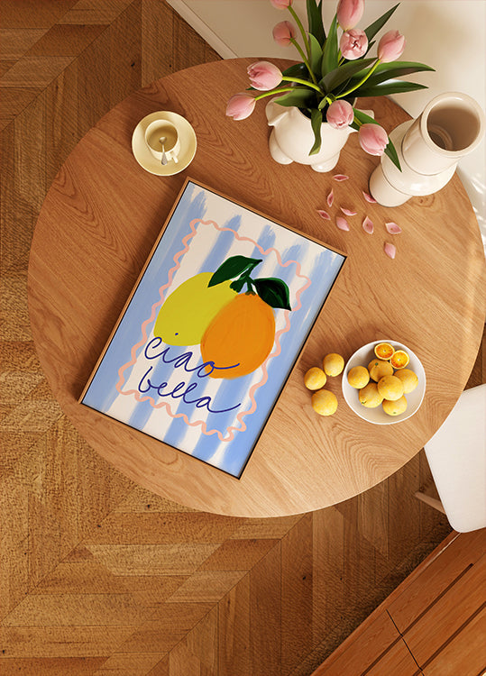 une table en bois avec une œuvre d'art encadrée représentant deux citrons, un bol de fruits et un vase de fleurs roses.