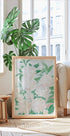 une grande impression encadrée de fleurs blanches avec des feuilles vertes, placée sur un parquet dans une pièce lumineuse et aérée avec de grandes fenêtres.
